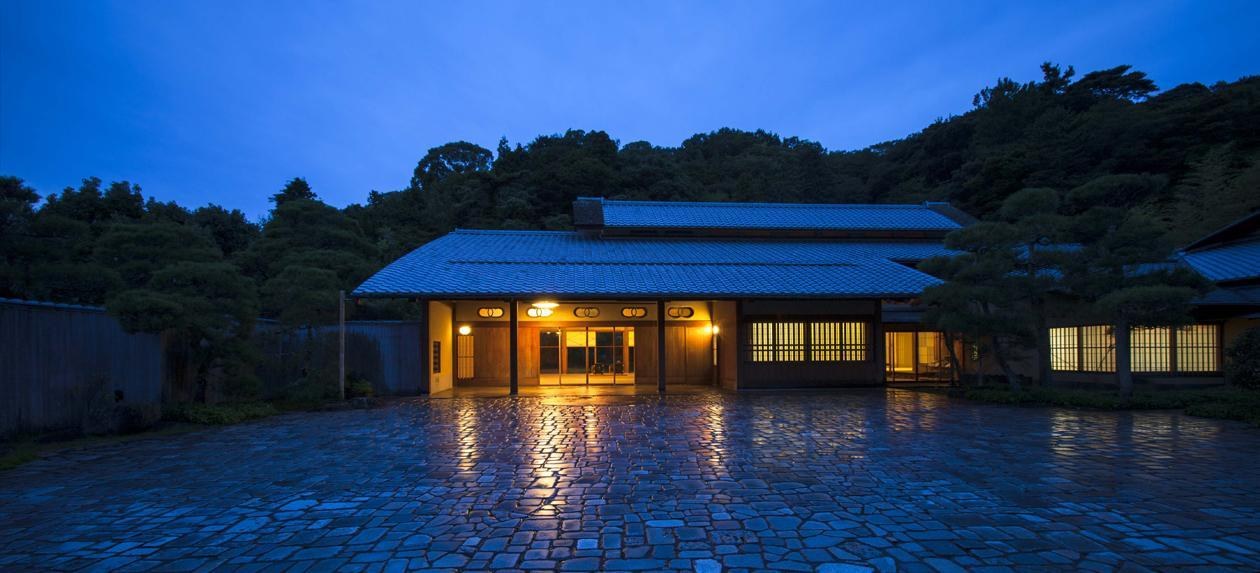 三養荘 — 伊豆の国市・夕景の玄関棟と石張りの前庭、瓦屋根の水平線と障子の灯りが石畳に映る