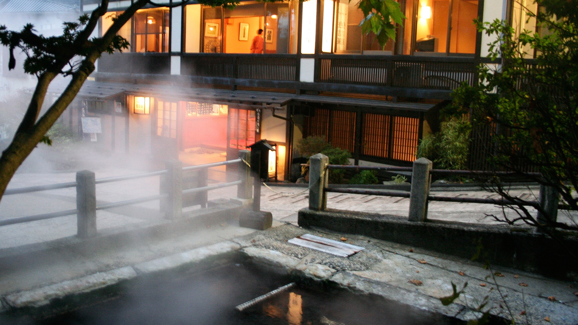 村のホテル 住吉屋 — 野沢温泉・麻釜前 · 湯けむりの上がる源泉・麻釜と、その縁に建つ木造三階建ての本館外観